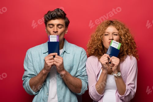 Preview: couple of happy travelers kissing passports with air tickets, isolated on red