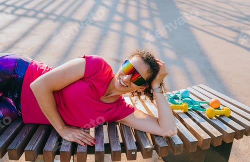 Preview: Smiling woman in sporty sunglasses relaxing on a bench after a workout, surrounded by dumbbells and