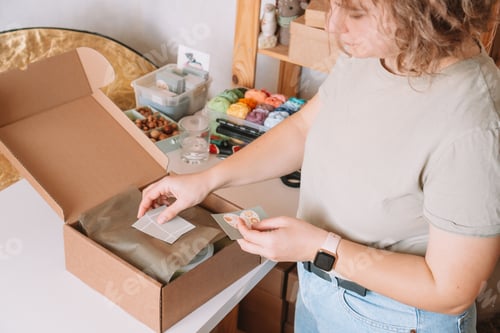 Preview: Smiling blond curly woman packing and wrapping shipment with postcards, silicone baby dishes in