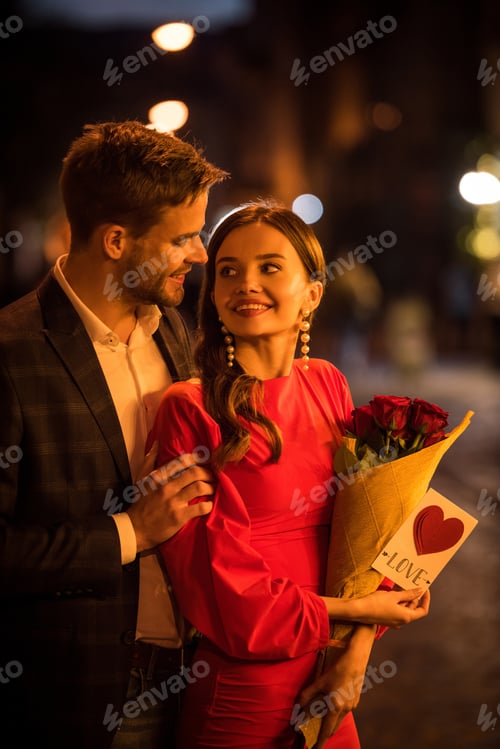 Preview: smiling man hugging happy girlfriend holding bouquet of roses and card with love inscription and