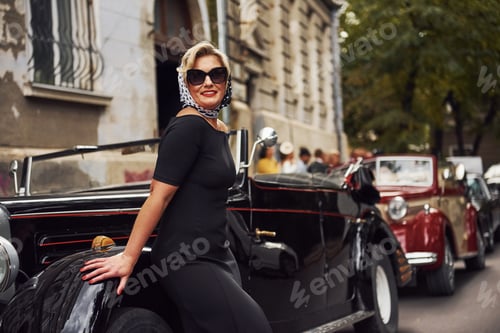 Preview: Blonde woman in sunglasses and in black dress leaning on old vintage classic car