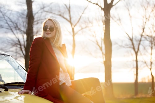 Preview: Style blonde woman in sunglasses and red coat sits on car at countryside