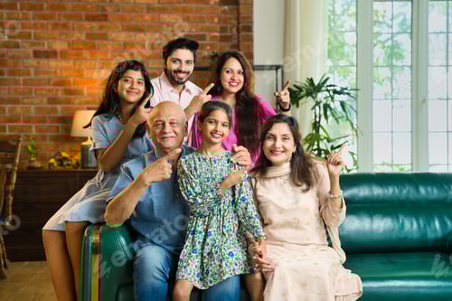 Preview: Indian multigenerational family of six sitting on a sofa, happily pointing to advertise something