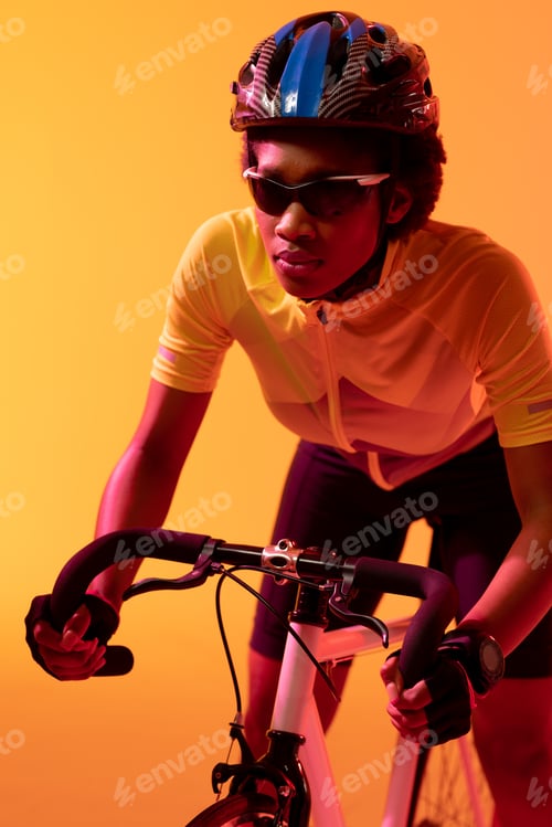 Preview: Vertical image of african american female cyclist riding bike in neon orange lighting