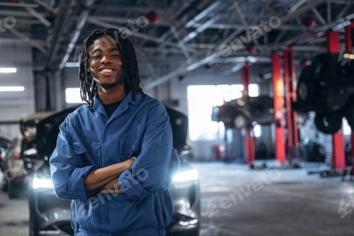 Preview: Friendly male mechanic is with car in garage, standing with arms crossed