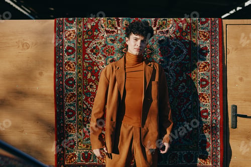 Preview: Stylish young man in a brown outfit standing against a patterned rug backdrop The warm tones create