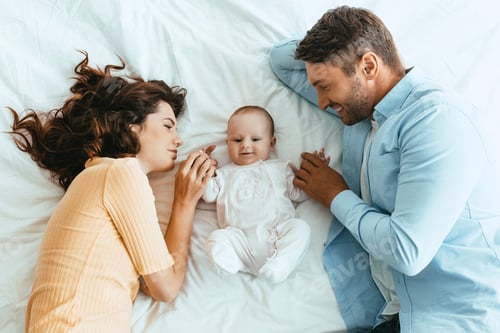 Vista previa: feliz mamá y papá tocando suavemente al adorable bebé mientras están acostados juntos en ropa de cama blanca