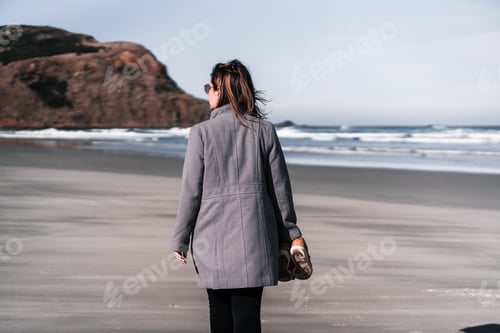 Preview: blonde caucasian girl in a long gray coat black pants sunglasses and with the slippers in her right