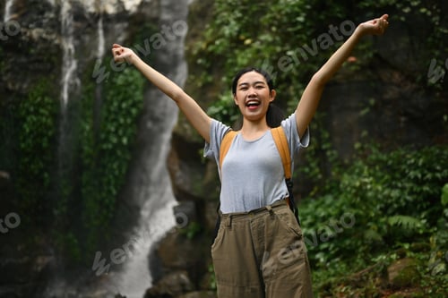 Preview: Cheerful female traveler raising arms enjoying the view of beautiful tropical waterfall