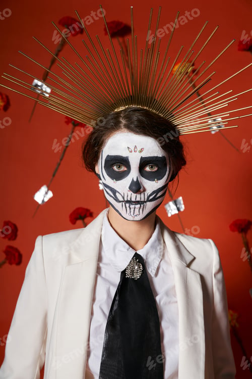 Preview: extravagant woman in skull makeup and white suit with crown looking at camera on red floral backdrop