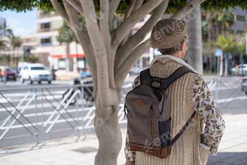 Preview: Rear view of caucasian mature woman with cap and backpack walking in the city street