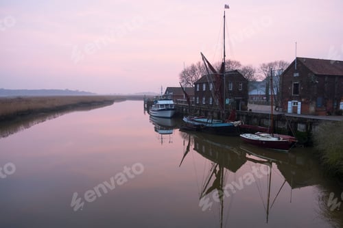 Preview: Sailing boats moored at harbour wall, reflected on the surface of a calm canal at twilight.