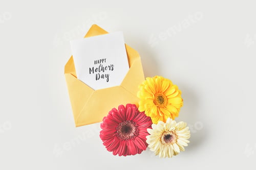 Preview: top view of happy mothers day greeting card in envelope and beautiful gerbera flowers on grey