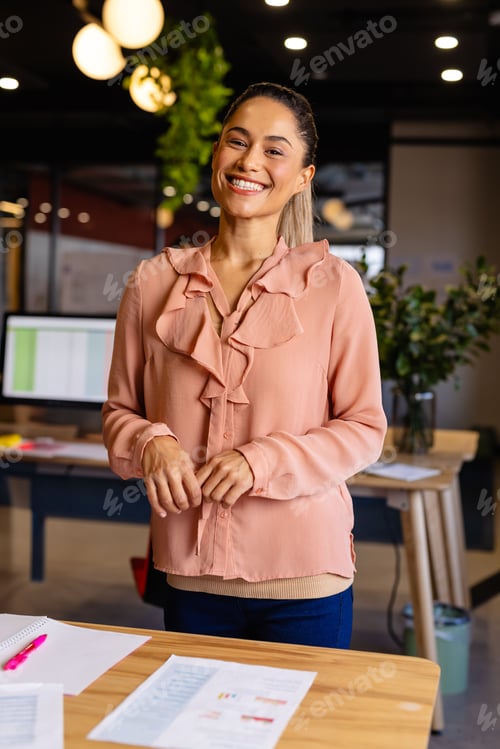 Preview: Woman in 30s standing behind wooden desk in modern office, smiling and reviewing charts