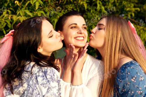 Preview: Charming young bride in a white dress smiles happily and two bridesmaids with pink veils kiss her on