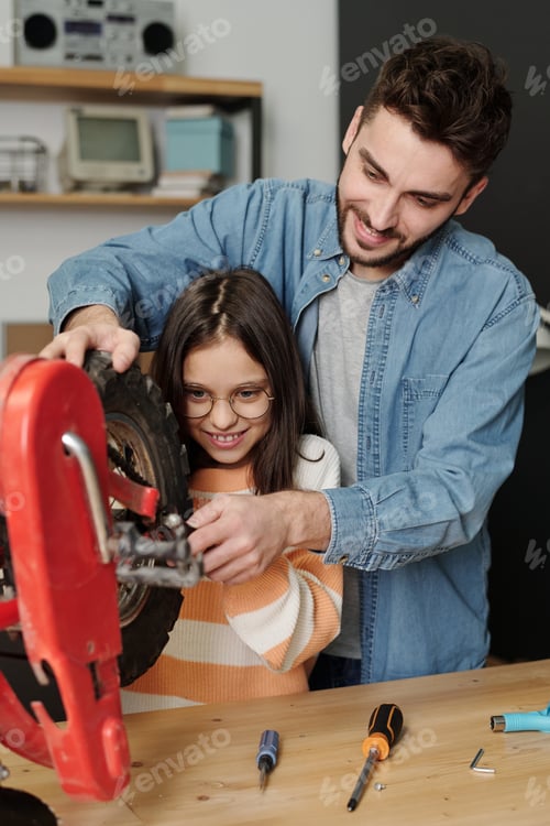Preview: Young man showing his little daughter how to fix part of bicycle wheel in garage
