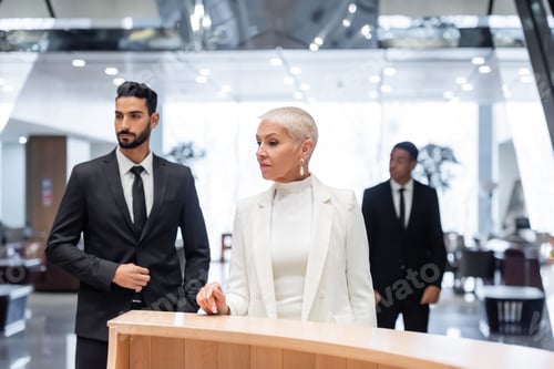Preview: wealthy business lady waiting at hotel reception near interracial bodyguards