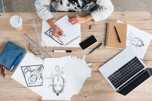 Preview: cropped image of female artist writing in textbook at table with laptop and smartphone