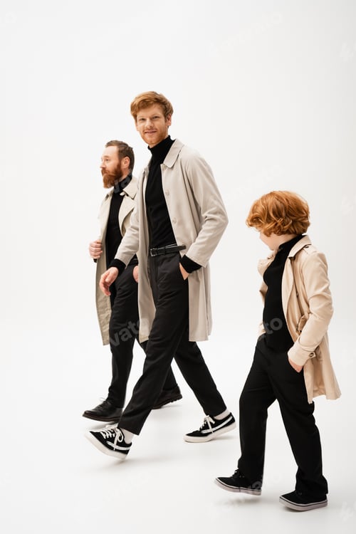 Preview: full length of trendy bearded men and redhead boy in trench coats walking on light grey background