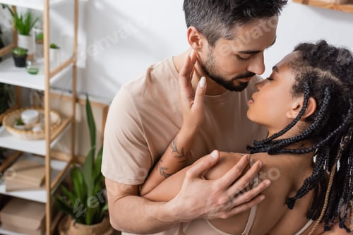 Preview: bearded brunette man in t-shirt hugging seductive african american woman with dreadlocks in bedroom