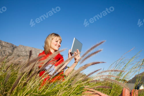 Preview: Young woman sitting in long grass using digital tablet