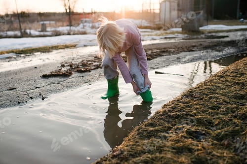 Preview: Boy rolling trousers up in rural meltwater puddle