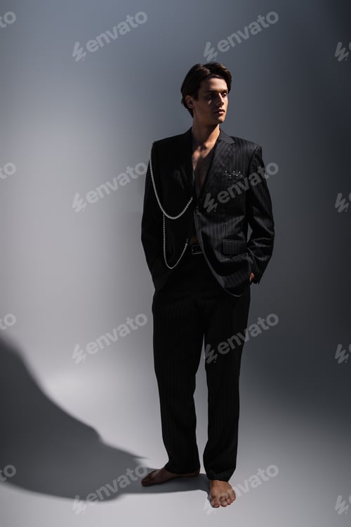 Preview: full length of man in trendy suit with pearls looking away while posing with hands in pockets on