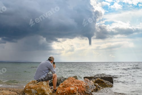 Preview: Man taking photo and filming waterspout or tornado in sea stormy weather with dramatic cloudy sky