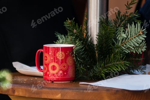 Preview: Closeup of the red Christmas cup with yellow patterns next to the pine tree branches on the table