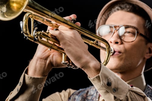 Preview: close up view of mixed race male jazzman in hat and eyeglasses playing on trumpet isolated on black