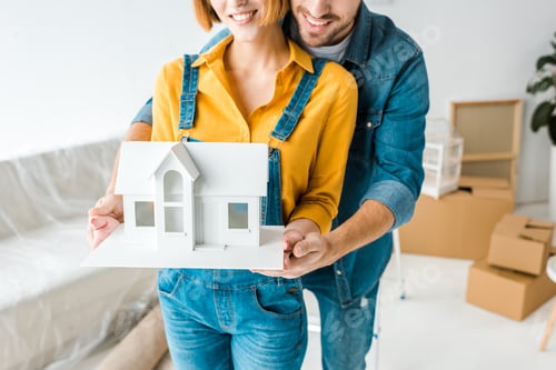 Preview: Cropped view of smiling couple holding toy house and looking at camera at home