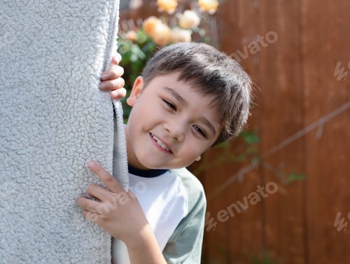 Preview: Happy kid playing hide and seek in the garden, boy with smiling face while standing behind blanket