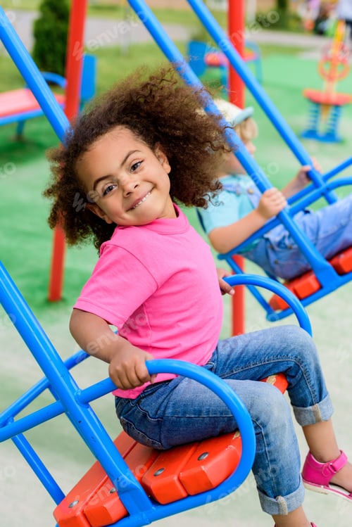 Preview: smiling curly african american little child riding on swing at playground