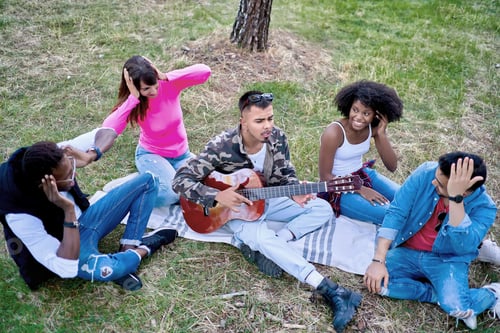 Preview: Friends sitting on the ground while one plays a horrible guitar in a park.