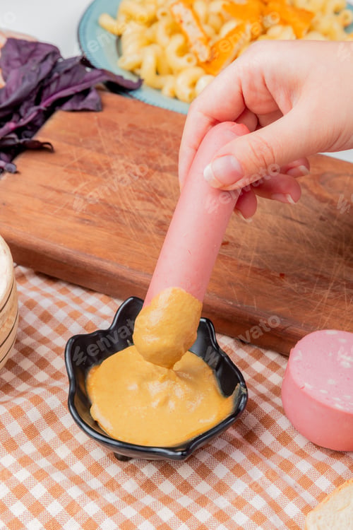 Preview: side view of female hand immersing milky sausage in bowl of mustard on plaid cloth
