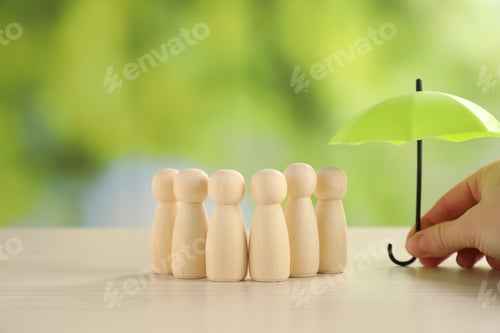 Preview: Woman holding umbrella over human figures at light wooden table outdoors, closeup. Insurance concept