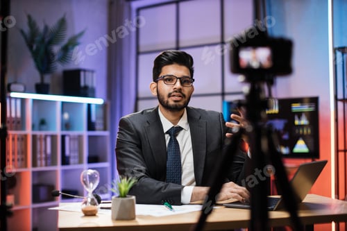 Preview: Confident indian bearded businessman in suit sitting in front of camera in evening office