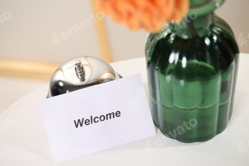 Preview: Card with word Welcome, desk bell and vase with flower on white table indoors, closeup