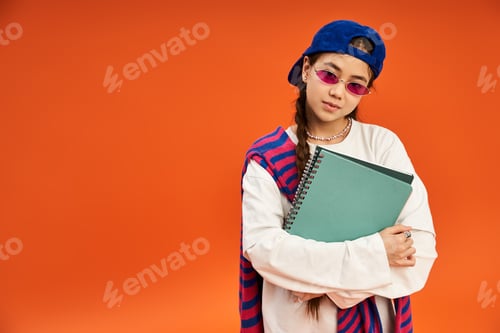 Preview: Stylish teenage girl poses in trendy outfit against vibrant orange backdrop in studio