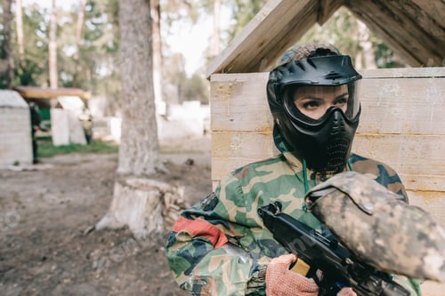 Preview: serious female paintballer in protective mask and camouflage uniform standing with paintball gun