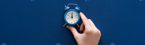 Preview: cropped view of woman holding small alarm clock on blue background, panoramic shot