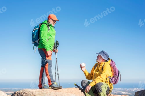 Preview: Happy elderly hikers friends with backpacks sit talking at the summit after a mountain hike