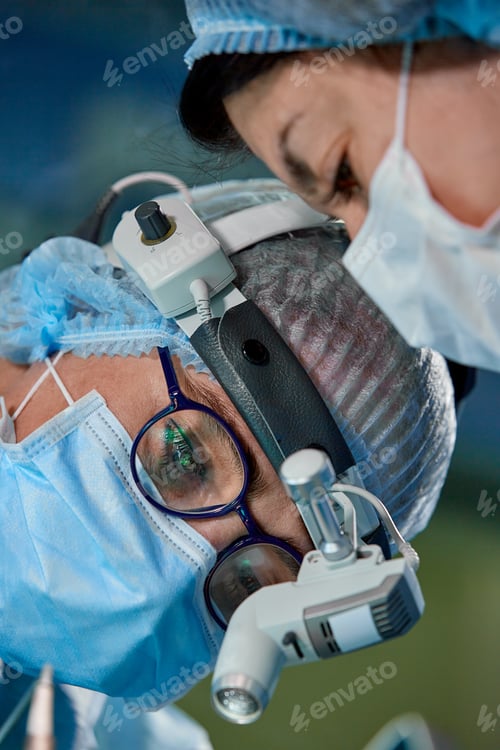 Preview: Surgeon performing cosmetic surgery in hospital operating room. Surgeon in mask wearing loupes