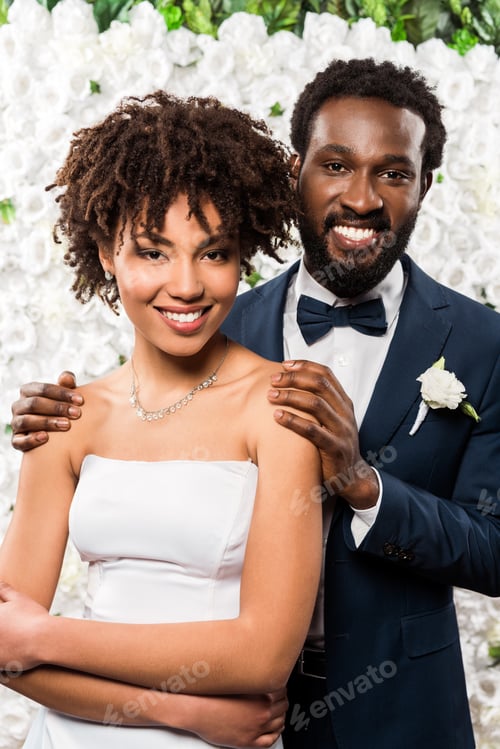 Preview: cheerful african american bridegroom and bride looking at camera near flowers