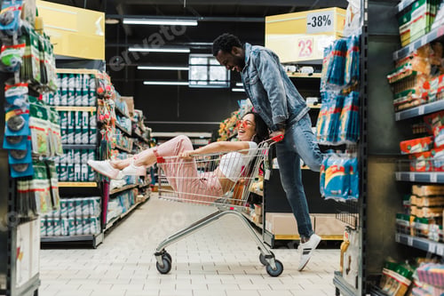 Preview: happy african american man looking at asian girl sitting in shopping cart in supermarket