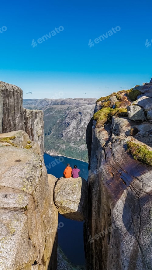 Preview: Two individuals sit on a narrow rock ledge overlooking a deep valley in NorwaynKjeragbolten cliff