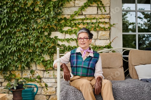 Preview: attractive joyous mature woman sitting on sofa near her house in England and looking at camera