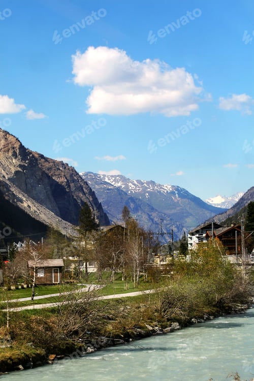 Preview: Mountain village with a river flowing under a blue sky, surrounded by snow-capped peaks. Zermatt