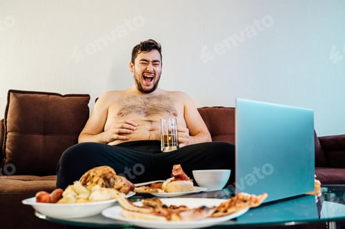 Preview: joyful fat man driking beer on the sofa and watching funny videos on laptop.