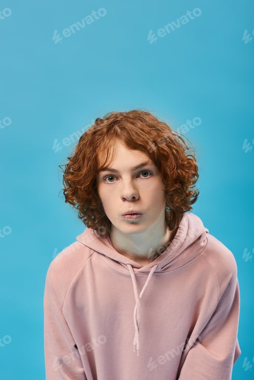 Preview: portrait of impressed teenage boy with red wavy hair in stylish hoodie and looking at camera on blue